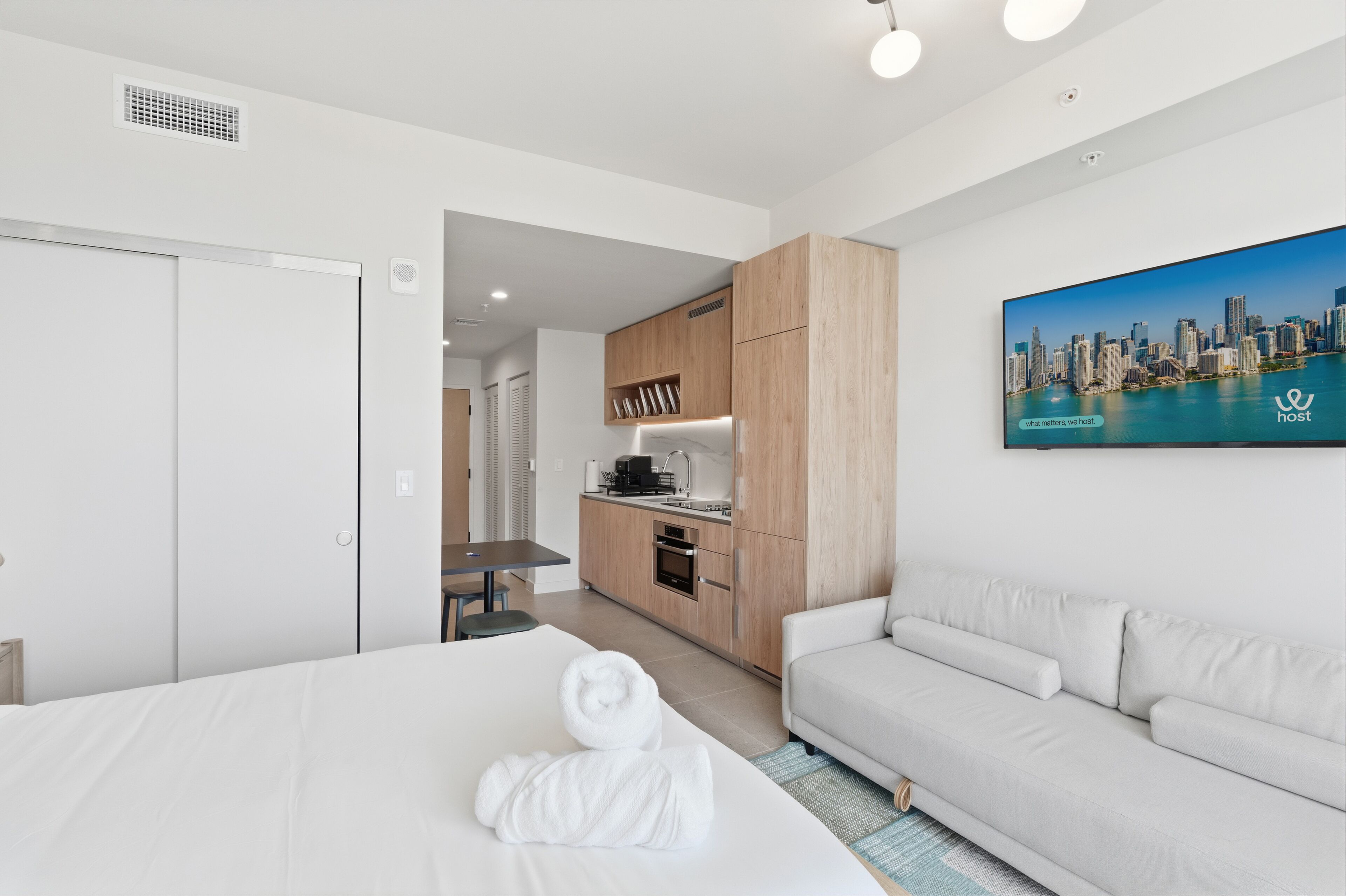 Une chambre d’hôtel avec un lit, un canapé blanc, une kitchenette et un tableau représentant un paysage urbain accroché au mur.