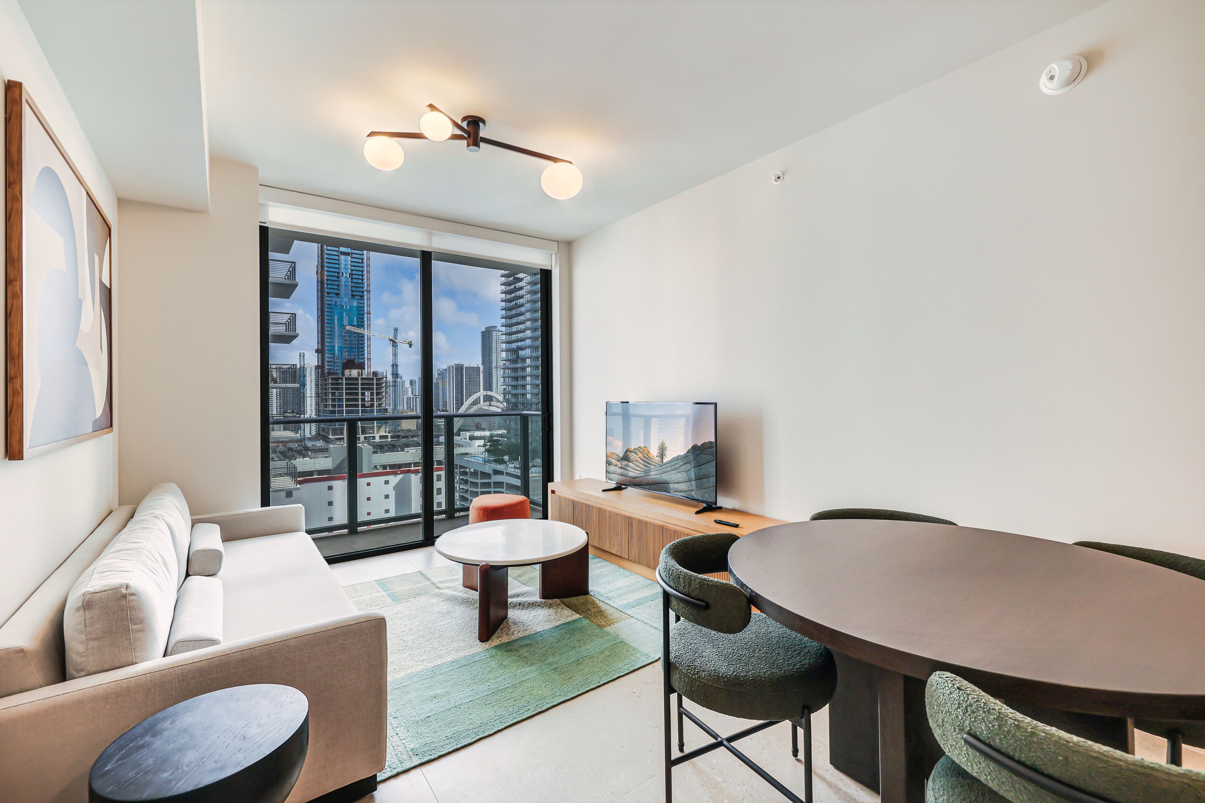A modern living room with a sofa, round coffee table, dining table with chairs, a television, and a city view through large windows.