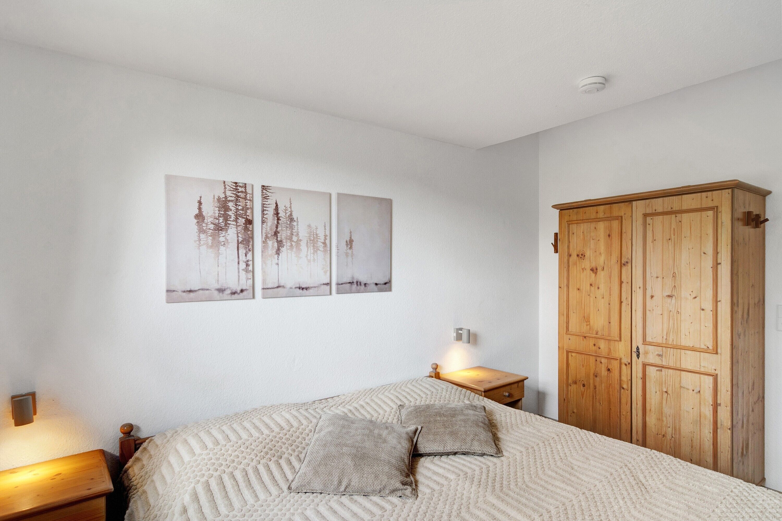 A bedroom with a bed, two bedside tables, a wardrobe, and three wall-mounted paintings.