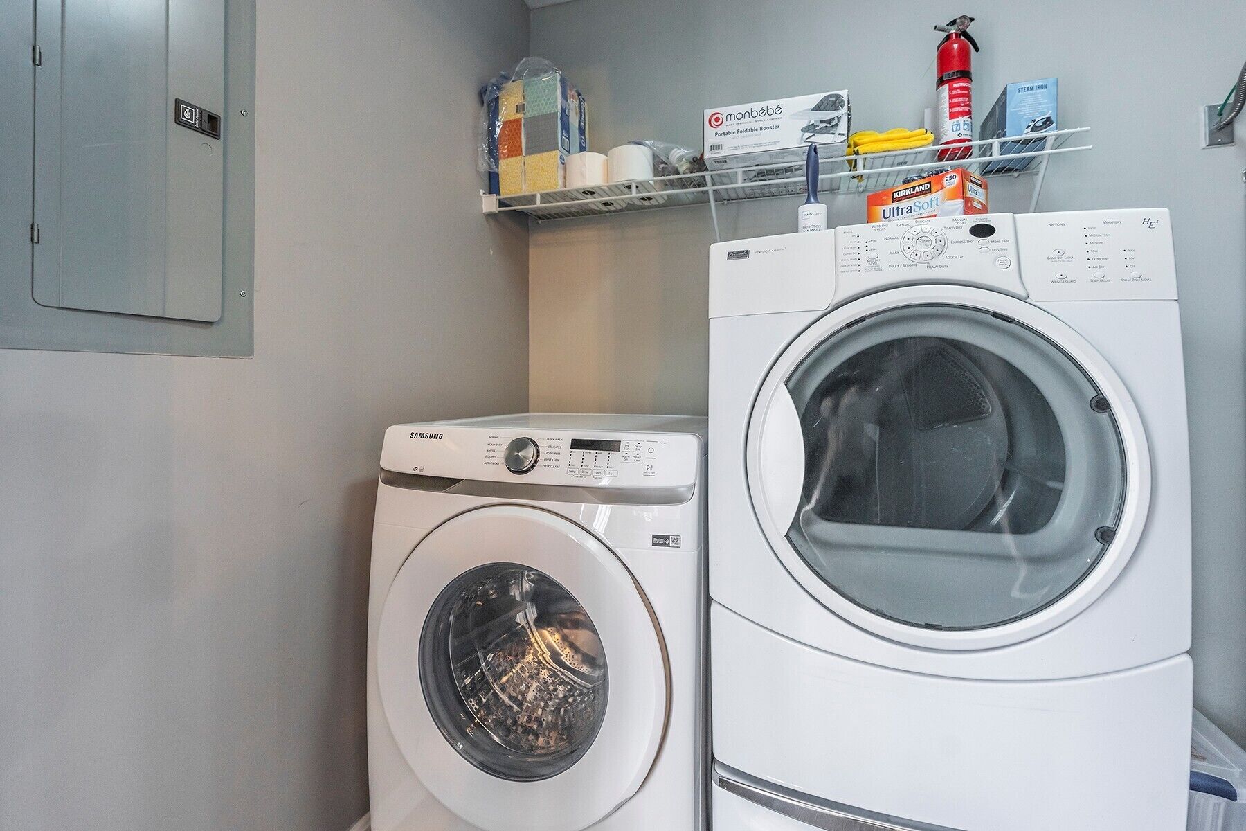 Deux machines à laver à chargement frontal, de couleur blanche, se trouvent dans un espace dédié à la buanderie. Une étagère située au-dessus contient divers objets.