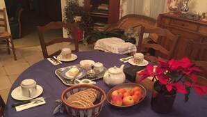 Une table à manger recouverte d’une nappe violette, un panier de pain, un saladier de pommes, une théière, des tasses et une plante en pot.