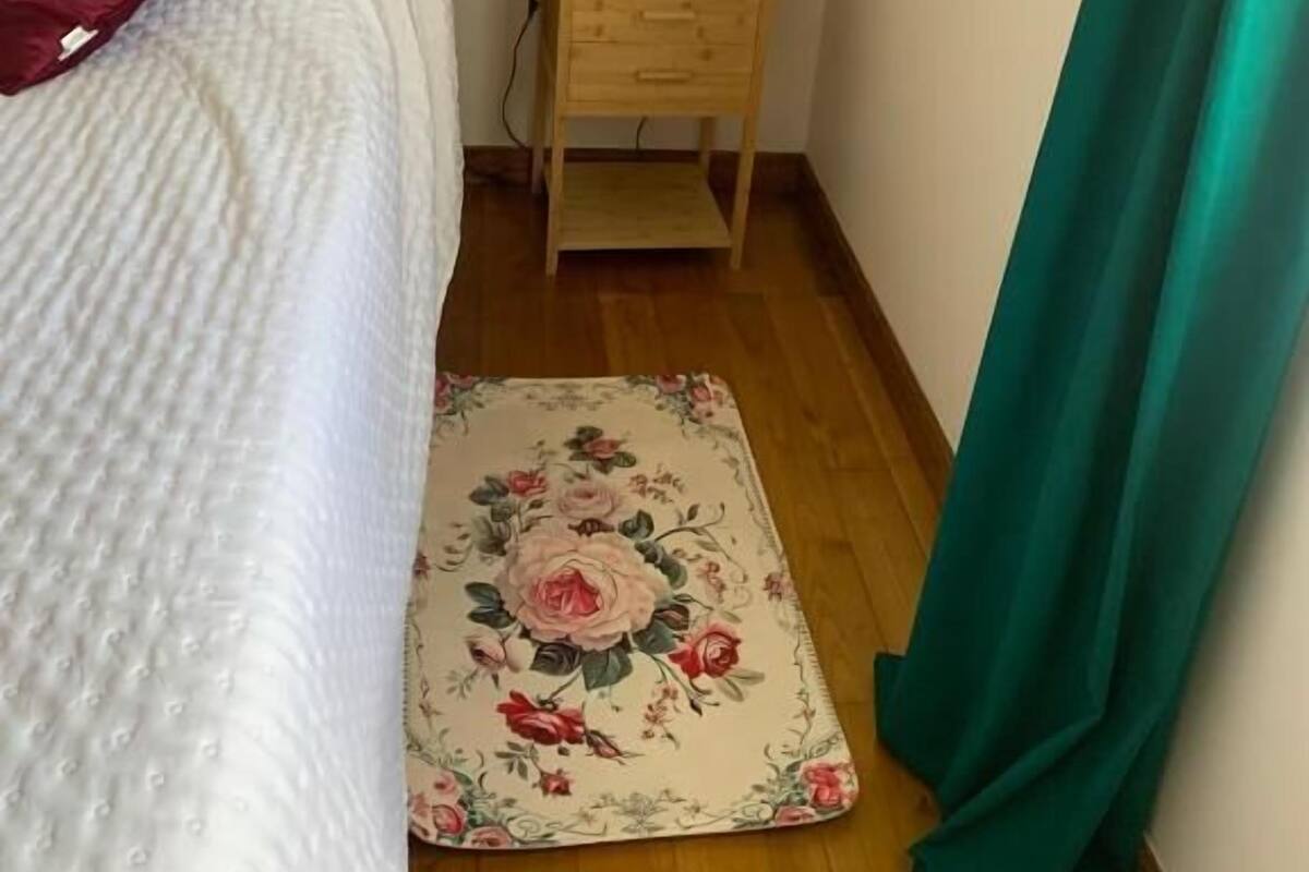 A bedroom with a bed, a nightstand with a lamp, a red box, and a floral rug.