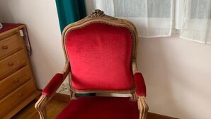 Un fauteuil rouge, recouvert de tissu, doté d’une structure en bois, placé dans une pièce dont le sol et la commode sont également en bois.