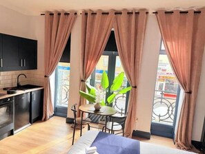 Una habitación de hotel compacta con kitchenette, zona de comedor y un amplio ventanal con cortinas.