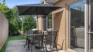 Un patio exterior con mesa y sillas, un paraguas grande y una puerta corrediza de vidrio que da a una zona de estar.