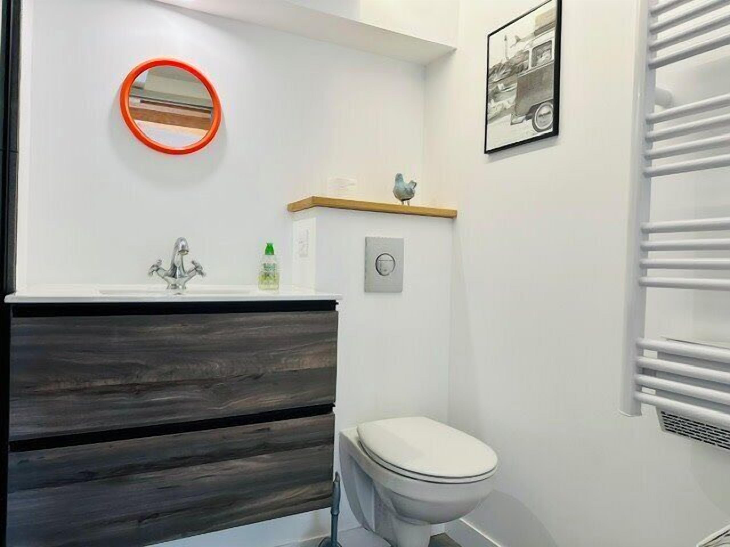 A bathroom with a white toilet, a wooden vanity, a mirror with an orange frame, a small shelf with a bird figurine, and a wall-mounted radiator.
