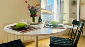 Ruokailutila, jossa on pyöreä pöytä, tuoleja, maljakko kukkineen ja ikkunasta avautuva näkymä rakennuksiin.