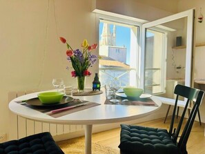 A dining area with a round table, chairs, a vase with flowers, and a view of buildings through a window. - Studio with Lantern Tower view near beach (La Rochelle)