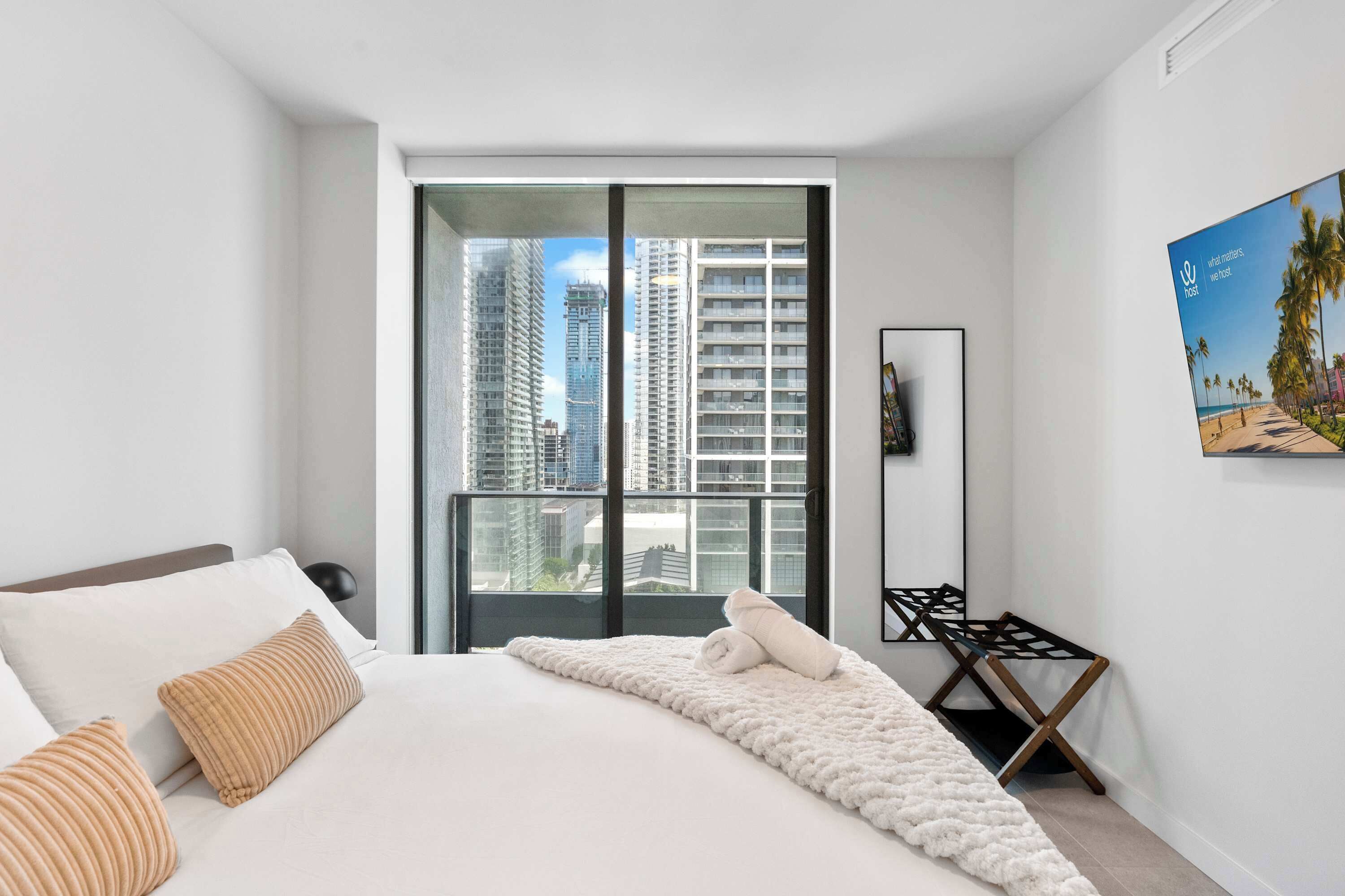 A modern hotel room with a large bed, a view of tall buildings, a black floor lamp, and a framed artwork on the wall.