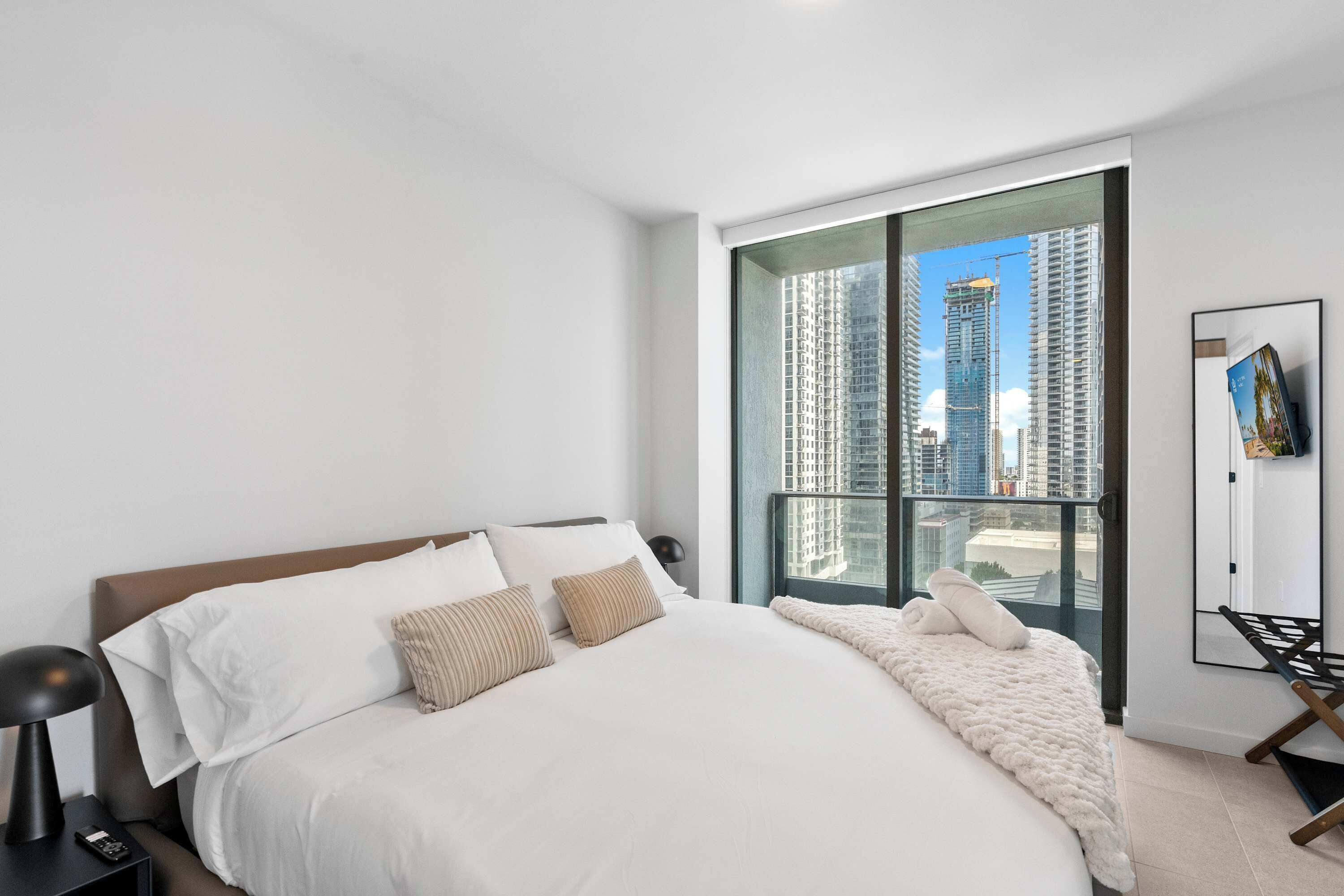 A modern bedroom with a large bed, a nightstand, a lamp, a window with a view of high-rise buildings, and a framed picture on the wall.