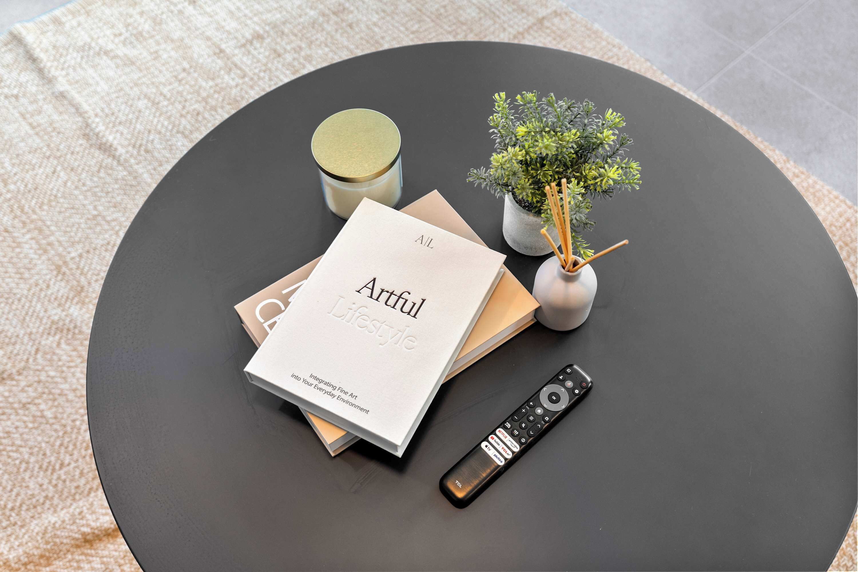 Una mesa redonda con un control remoto, un libro titulado 'Artful Lifestyle', una pequeña planta en maceta y una vela.