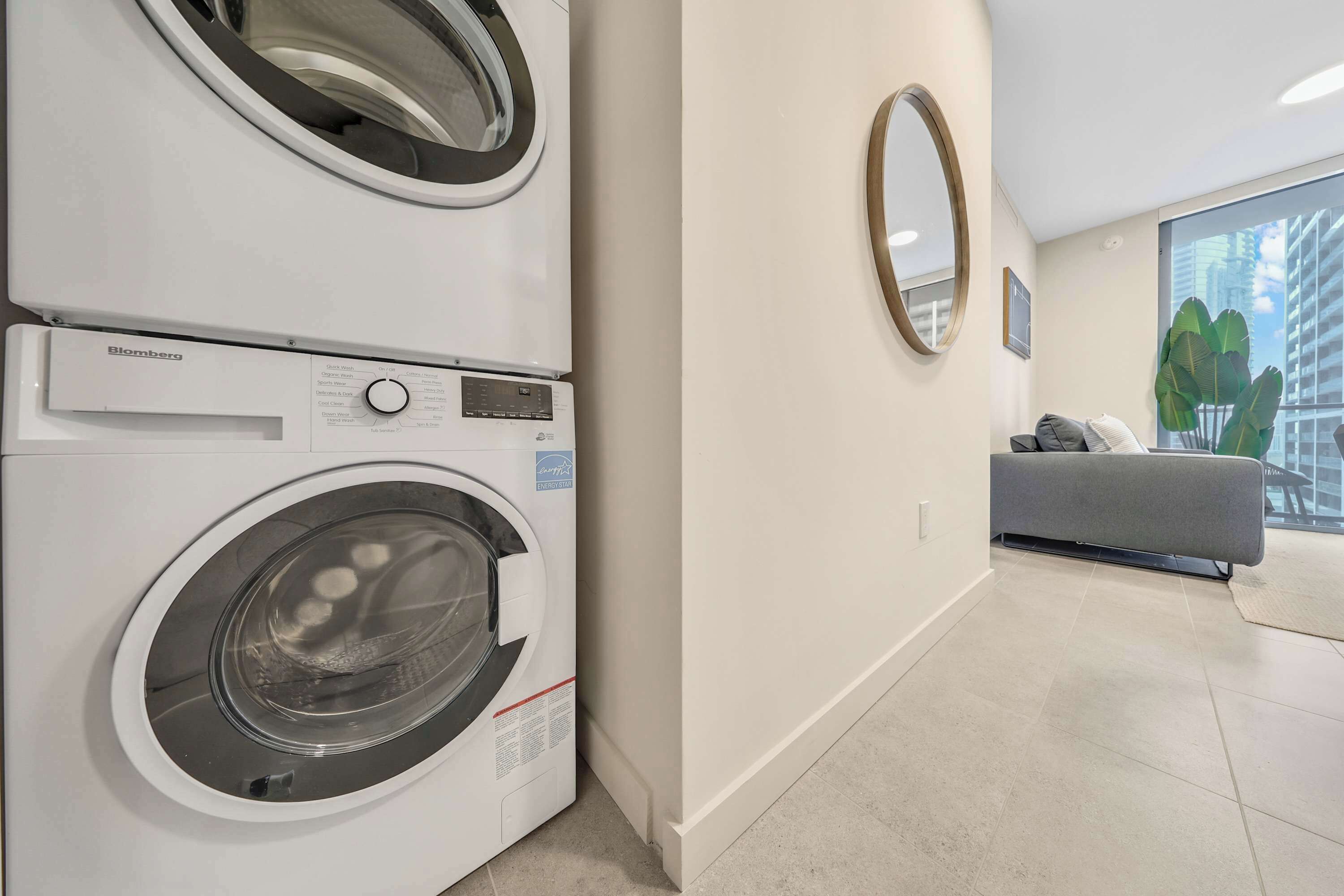 A modern laundry room with a front-loading washer and dryer, a gray sofa, a large mirror, and a view of tall buildings through a window.
