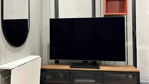 Egy modern nappali, melyben egy síkképernyős televízió, egy fa pad, egy fehér szekrény és egy nagy tükör található.