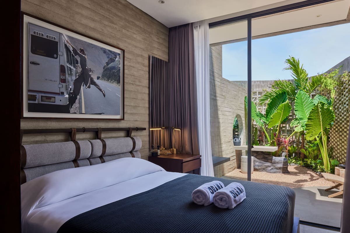 Ein Hotelzimmer mit einem großen Bett, einem Nachttisch, einer Lampe, einem Fenster mit Vorhängen und Blick ins Freie.