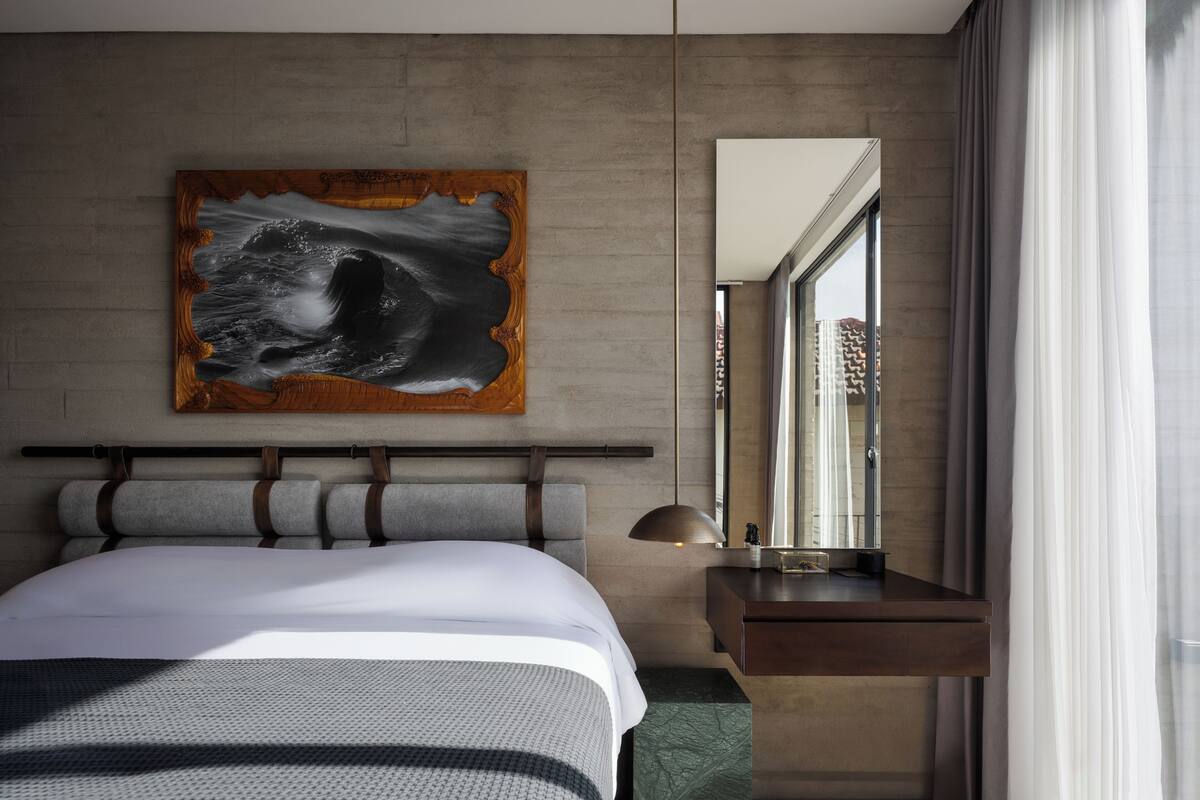 Ein modernes Hotelzimmer mit einem großen Bett, einem Holzkopfende, einem Gemälde, einem Spiegel, einem Nachttisch und Blick auf die Stadt durch das Fenster.