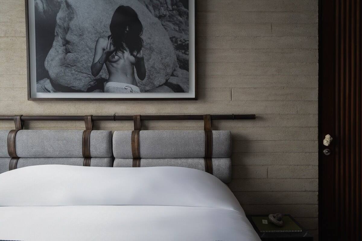 Ein Bett mit weißer Matratze, ein Kopfteil mit braunen Lederriemen, ein gerahmtes Schwarz-Weiß-Foto an der Wand und ein kleiner Nachttisch mit einem Buch und einem dekorativen Gegenstand.