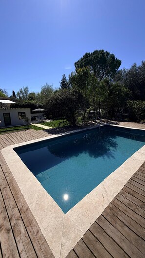 A rectangular swimming pool with a wooden deck surrounding it, set in a landscaped area with trees and a small building in the background. - Mazet Noa - Quiet terrace & pool (Le Rouret)