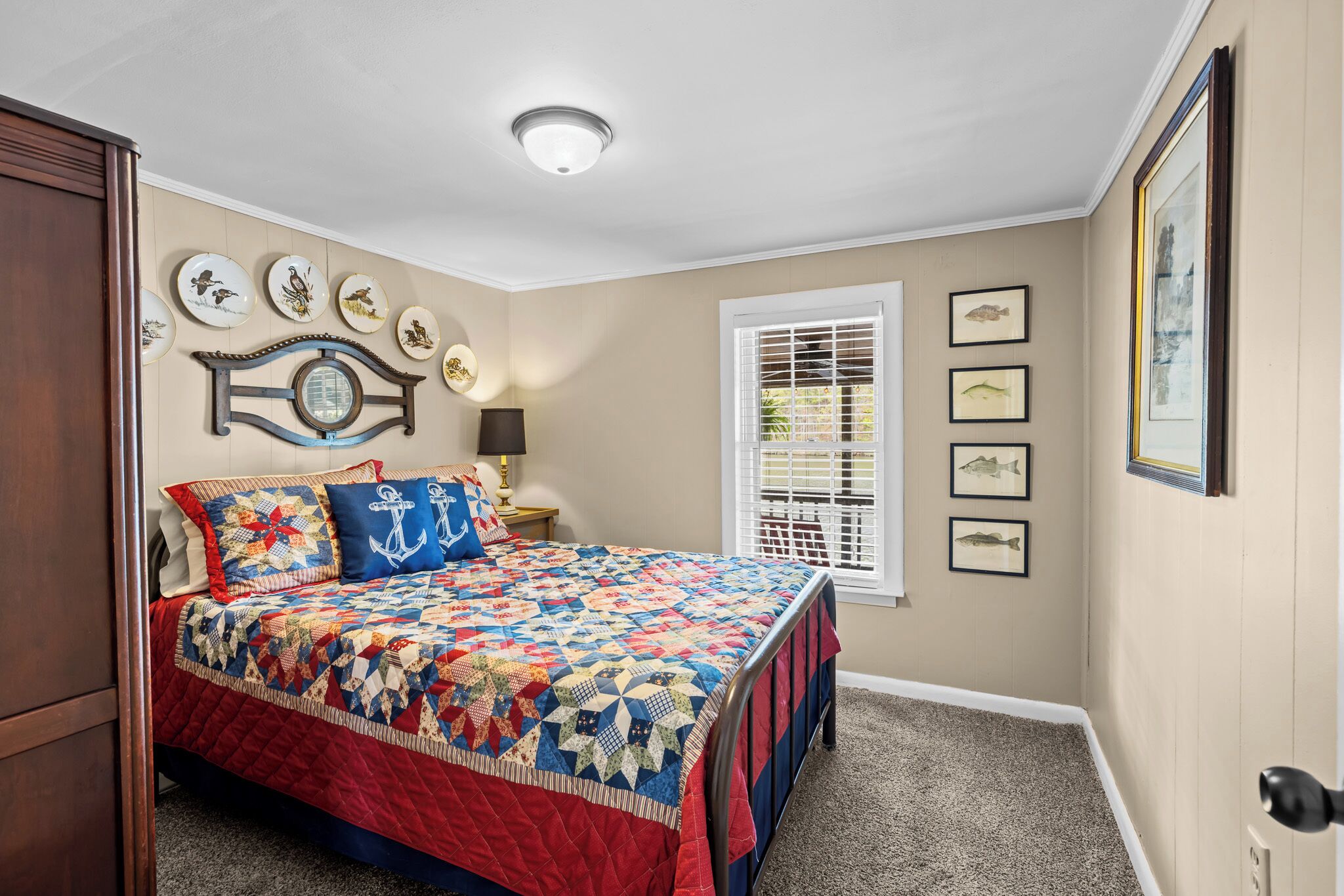 A bedroom with a large bed, a window with blinds, a wall clock, and framed pictures.