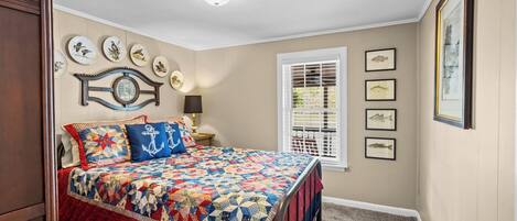 A bedroom with a large bed, a window with blinds, a wall clock, and framed pictures.