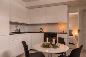 A modern kitchen with white cabinetry, a round dining table with candles, and a cozy living area visible through an open door. - Christmas in Stockholm, 20 min to T-Centralen (Hägersten)