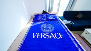 Une chambre d’hôtel avec un lit recouvert d’une couverture bleue ornée du logo « VERSACE », un canapé bleu foncé et une fenêtre avec des rideaux.