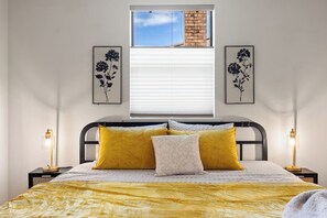 A bedroom with a bed, two bedside lamps, a window with blinds, and two framed floral artworks on the wall.