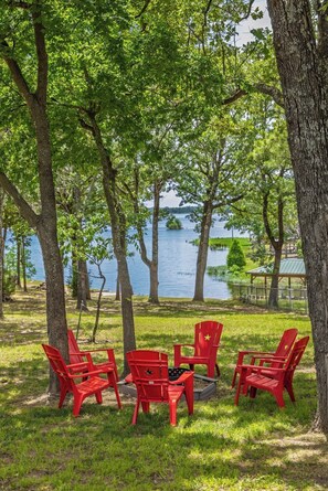 A lakeside outdoor seating area with red chairs and a fire pit.