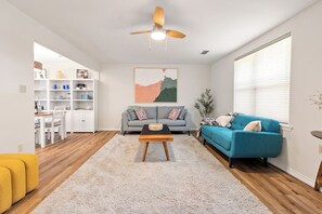 Una sala de estar con sofá, mesa de centro y ventilador de techo. Hay un comedor con mesa y sillas, y una cocina con armarios y estanterías.
