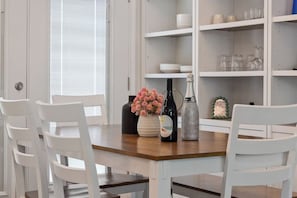 Un comedor con una mesa de madera, sillas blancas, un jarrón de flores, una botella y una jarra de cristal.