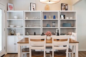 A dining area with a wooden table and white chairs, surrounded by white shelving units with various items.