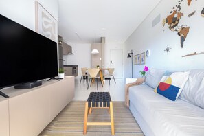 Uma sala de estar moderna com uma TV de tela plana, um banquinho de madeira, um sofá com almofadas coloridas, uma área de jantar com cadeiras e uma cozinha ao fundo.
