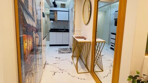 Un pasillo con suelo de mármol, una obra de arte enmarcada a la izquierda y un espejo redondo a la derecha, con una cocina al fondo.