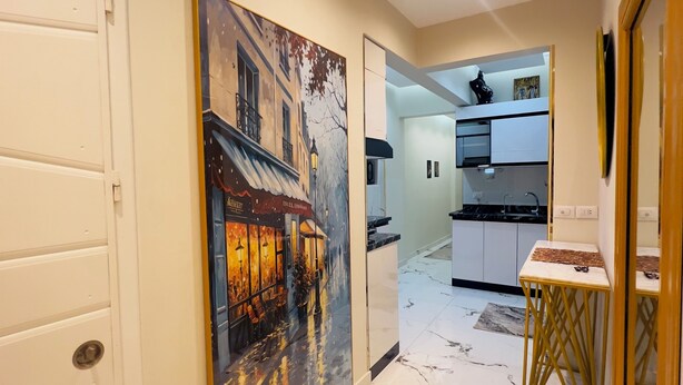 Un pasillo con piso de mármol, un cuadro de una escena callejera, una zona de cocina con armarios y un lavamanos, y una puerta con picaporte.