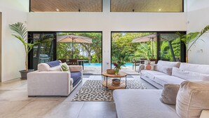 Amplia sala de estar con sofá, sillón y mesa de centro, una ventana grande y vista a una piscina y vegetación.