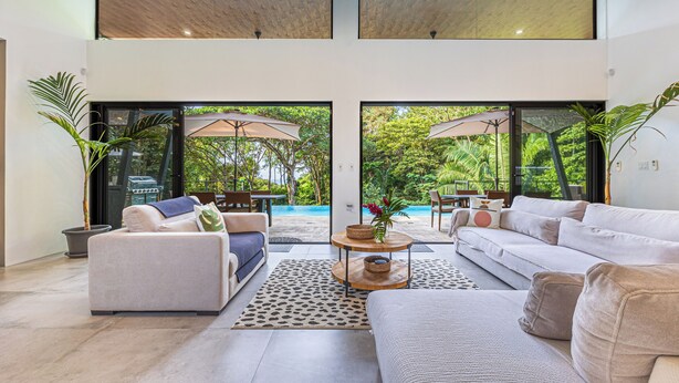 A spacious living area with a sofa, armchair, and coffee table, a large window, and a view of a pool and greenery. - Stunning 4-bedroom villa with infinity pool, 15 min. from Dominical (Uvita)