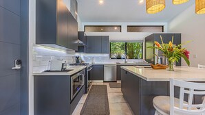 A modern kitchen with dark cabinetry, a central island, a built-in oven, and a window providing a view of greenery. - Stunning 4-bedroom villa with infinity pool, 15 min. from Dominical (Uvita)