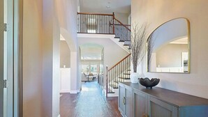 A spacious hallway with a wooden staircase, a large mirror, and a decorative vase on a cabinet. - 4BR Frisco Escape / Pool + Theater / Sleeps 10 / Spacious (Frisco)