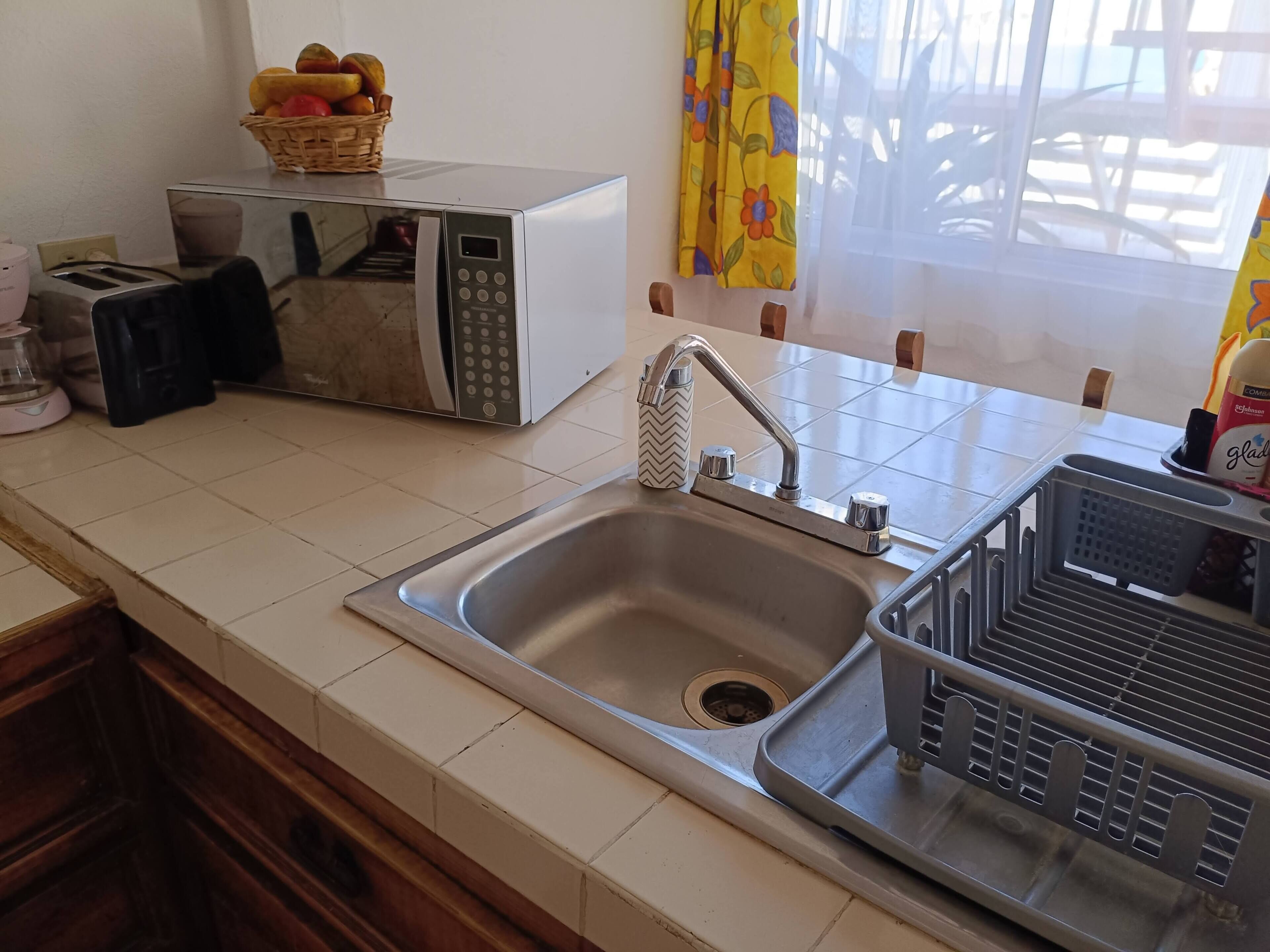 A kitchen with a stainless steel sink, a toaster, a microwave, and a basket of fruit on the counter.