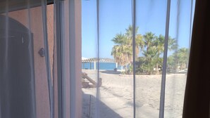 Vista a través de una ventana con persianas, mostrando una playa arenosa, palmeras y una estructura distante.