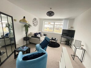A living room with a sofa, a coffee table, a TV, a lamp, and a large window. - Sapphire 1BR House Pet Friendly NR Addenbrookes (Cambridge)