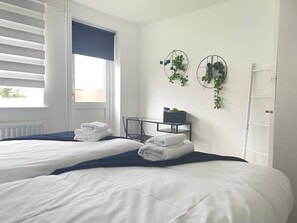 Un dormitorio con dos camas, una ventana con persianas, una mesita de noche con una planta y una silla.