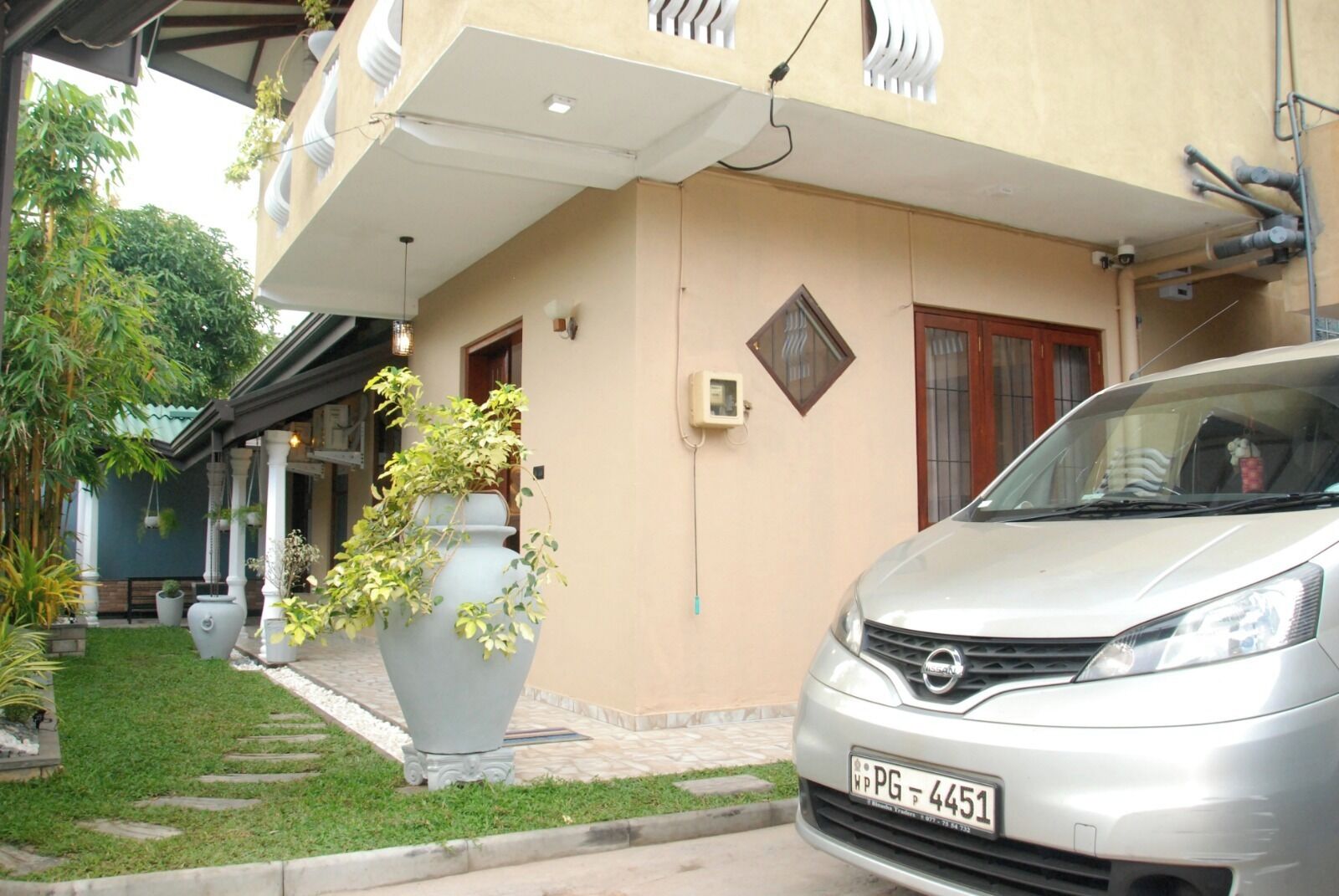 A residential building with a car parked in front, a large grey planter with plants, and a small window with a security camera.