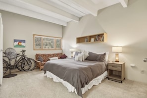 Een slaapkamer met een groot bed, een nachtkastje met een lamp, een boekenplank, een fiets en een schilderij aan de muur.