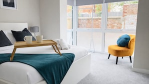 A modern hotel room with a bed, a small table with cups, a yellow armchair, and a large window with a view of a brick wall. - Amber 1BR Apart 14 min Walk to Tattersalls (Newmarket)