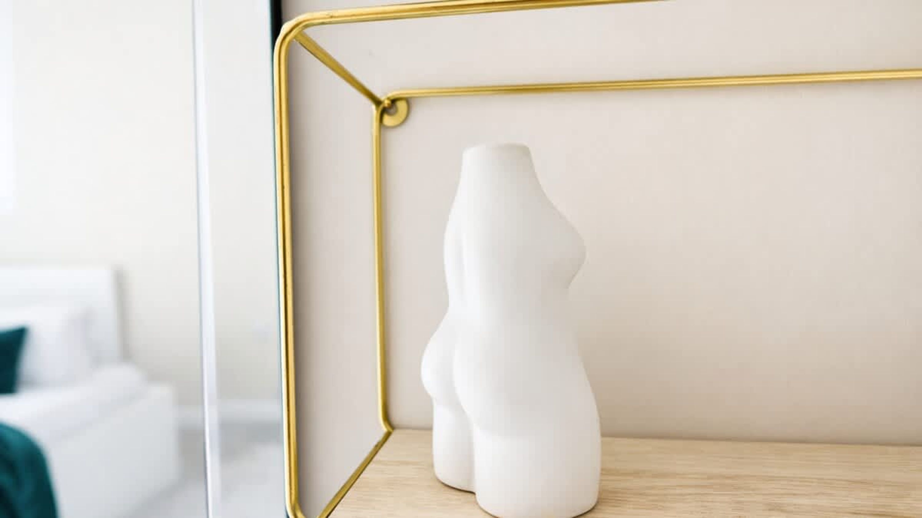 A white vase on a wooden shelf within a room with a white wall and a visible window.