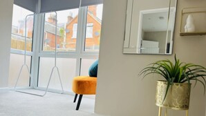 A room with large windows, a yellow chair, a plant on a stand, and a mirror on the wall. - Amber 1BR Apart 14 min Walk to Tattersalls (Newmarket)