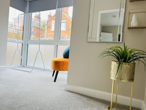 A room with large windows, a yellow chair, a plant on a stand, and a mirror on the wall. - Amber 1BR Apart 14 min Walk to Tattersalls (Newmarket)