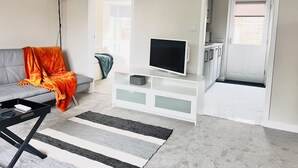 A living room with a grey sofa, a TV on a white stand, a black coffee table, a striped rug, and a door leading to a kitchen. - Amber 1BR Apart 14 min Walk to Tattersalls (Newmarket)