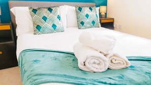 Un letto rifatto con cura, coperto da una trapunta color petrolio e decorato con due cuscini a motivi, un asciugamano bianco piegato e un comodino con una lampada.