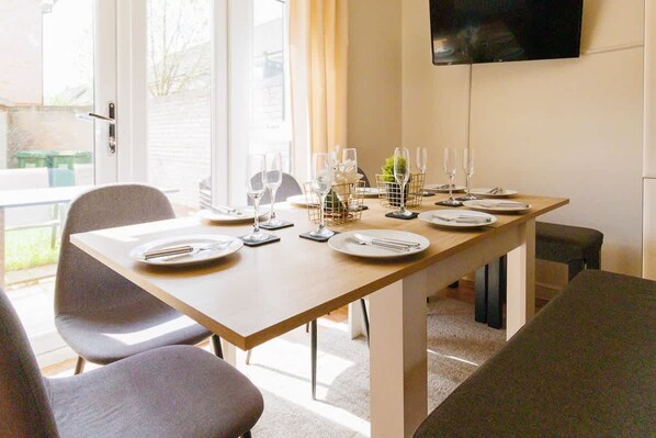 Une table à manger est dressée avec des assiettes, des couverts et des verres. Un téléviseur est fixé au mur et une porte donne sur l’extérieur.